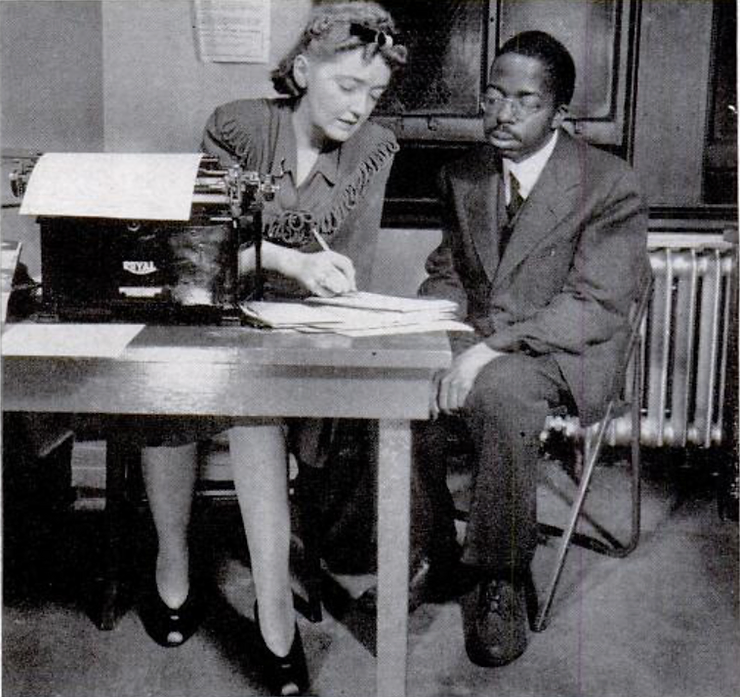 Photo from of Frances Sweeney at her desk taking notes from a fellow black editor, William Harrison, sitting beside her.
