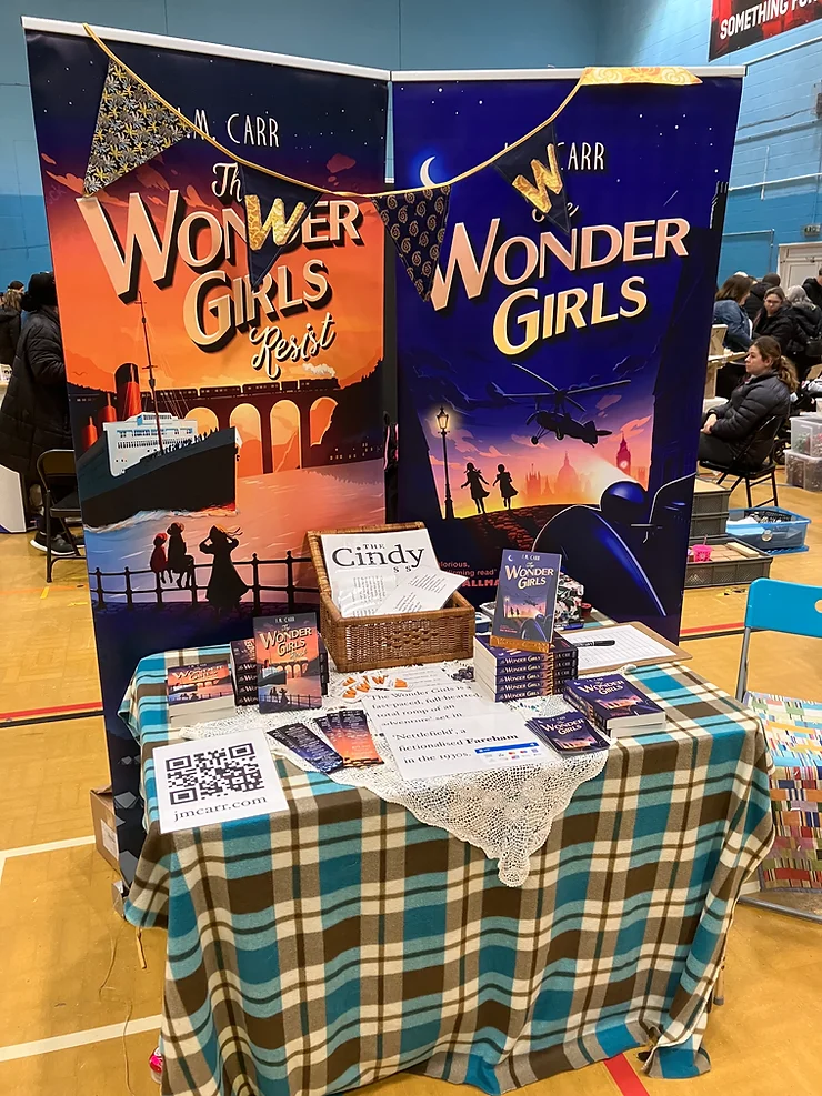 A photo of my stall at my first craft Market in Fareham. 2 big freestanding banners behind a plaid blanket covered table on which are  2 piles of books and some signage about the books.